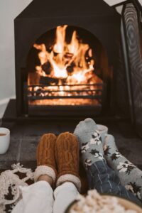 There is a fire in the background with two pairs of feet in the foreground. Each pair of feet has a pair of socks on. The pair on the left is tan and knitted, the pair on the right is gray with white flowers. They are practicing hygge together.
