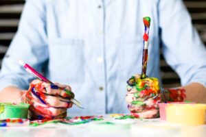 a person in a blue shirt sits at a desk covered in paint. their hands are also covered in paint, both holding paint brushes.
