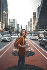 a woman with a backpack on stands on a bike path in the middle of a busy road, looking towards the camera, smiling. 