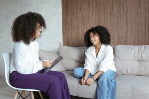 Woman in White Long Sleeve Shirt and Blue Denim Jeans Sitting on Gray Couch talking about chronic stress management