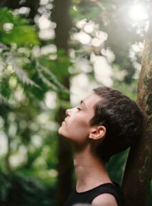A person with short hair leaning against a tree, their head tilted upward. They are practicing mindfulness to help maintain their motivation, as they learned through working with therapists at Awakened Path Counseling in New Jersey.