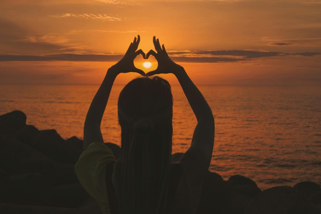 woman forming her hands in a heart in front of the sunset, representing self-compassion