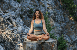A woman sits cross-legged on a large tree stump in the mountains, meditating with her eyes closed and hands resting on her knees. This serene scene reflects nature-based grounding practices often used in energetic hygiene to release stress and reconnect with the body.