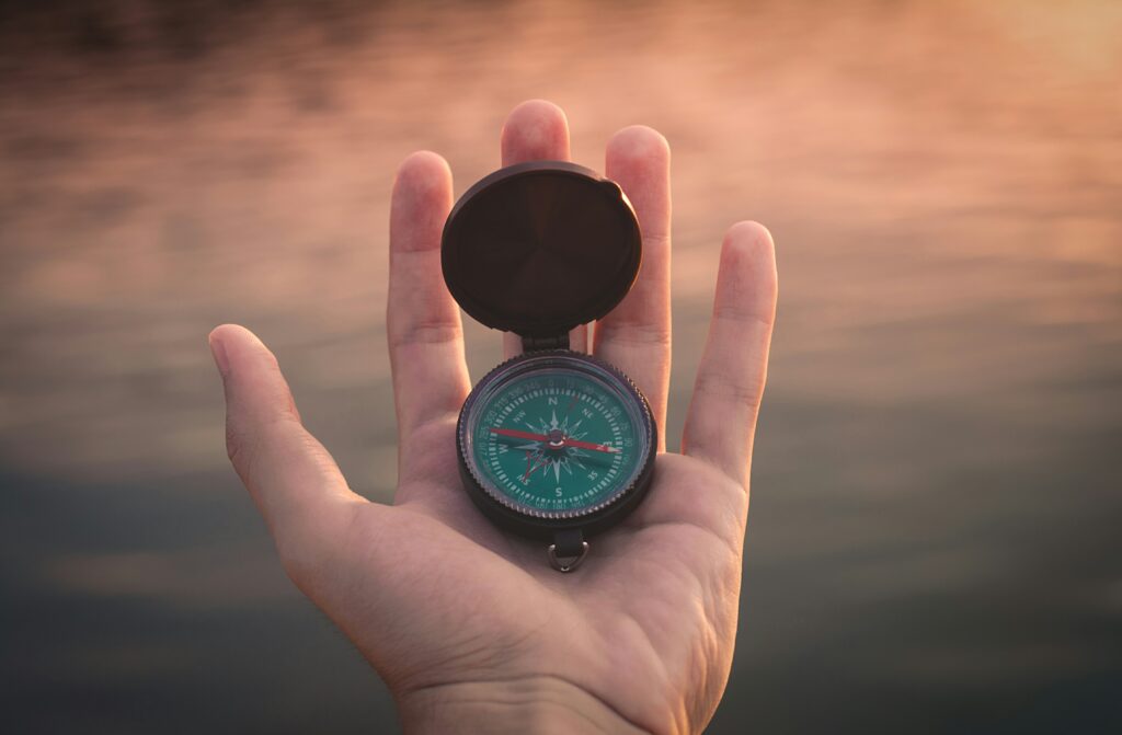 Hand holding an open compass, with the needle pointing north, set against a soft, sunset-colored background near the water, symbolizing guidance and direction.
