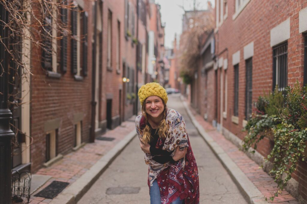 Woman standing in a narrow brick alley smiling warmly, representing the comfort, connection, and emotional regulation fictional stories can provide.