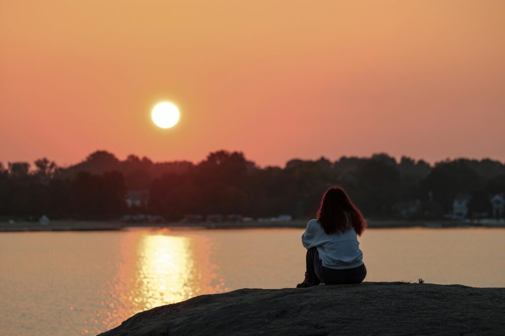 Person sitting silently by a lake at sunset, demonstrating mindful presence and somatic therapy techniques for stress reduction and emotional well-being.