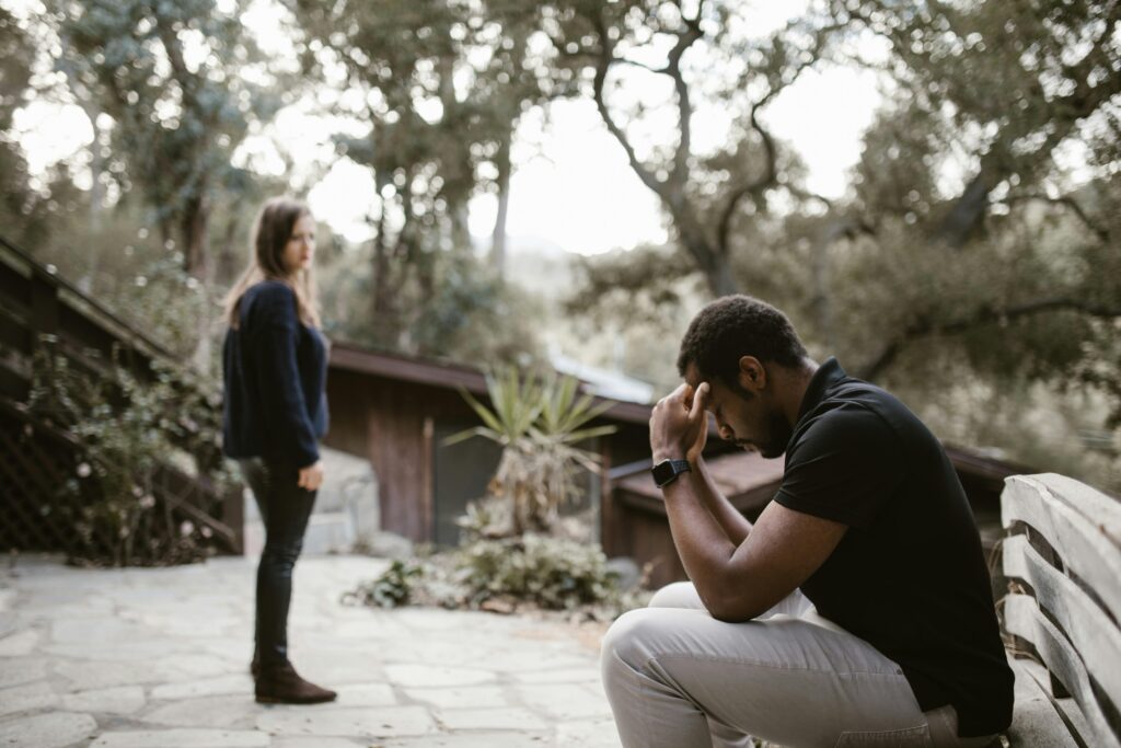 Man sitting with head in hands while a partner stands nearby, illustrating the emotional pain, confusion, and isolation of betrayal trauma.