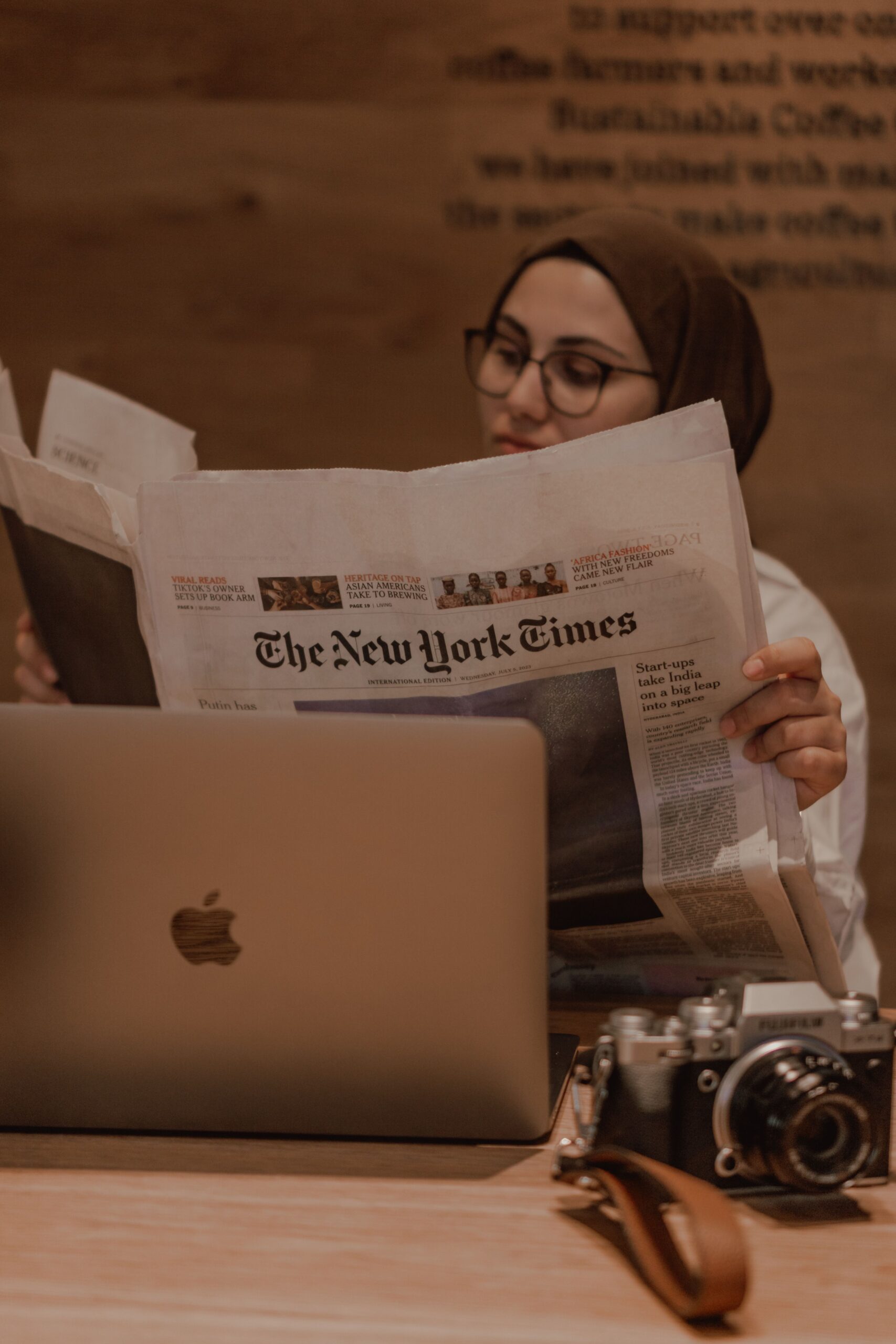 Woman reading a newspaper beside a laptop, illustrating information overload, constant news consumption, and anxiety in a digital world