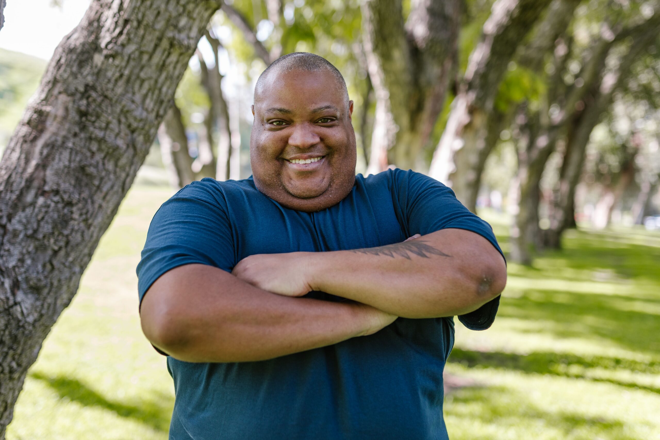 Smiling man standing with his arms crossed in a sunny park, representing resilience and support during emotional exhaustion.